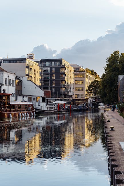 A canal bordered by a paved walkway on the right side with a row of bollards visible along the edge. On the left side of the canal, several boats and houseboats are moored, including a white boat with