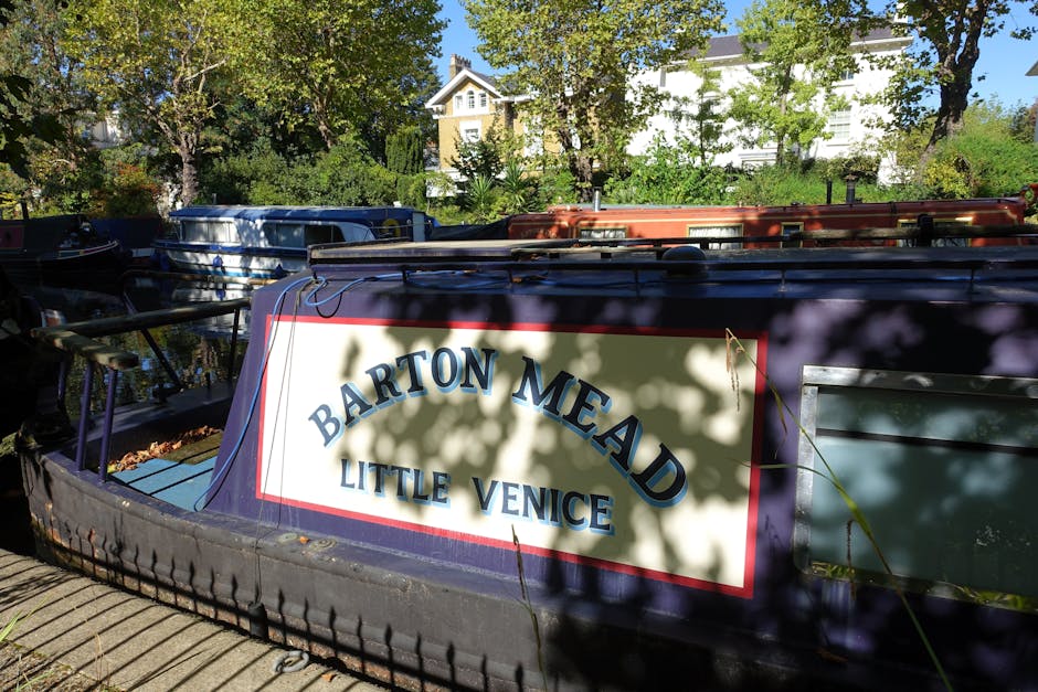 A canal bordered by a paved walkway on the right side with a row of bollards visible along the edge. On the left side of the canal, several boats and houseboats are moored, including a white boat with