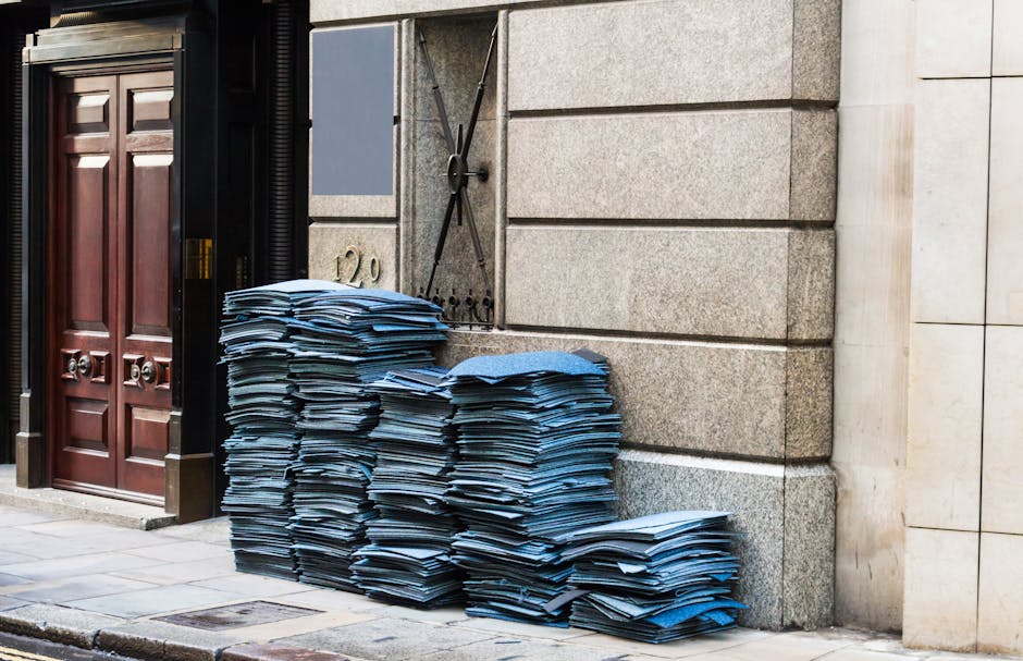 Two large black and white plastic rubbish bags are positioned on a paved sidewalk in front of a black metal fence with vertical bars. The bags appear to be filled with waste material and are slightly