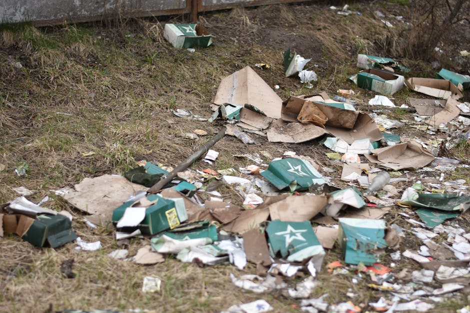 A small, cluttered area at the edge of a natural water body shows various pieces of mixed waste debris, including plastic bottles, packaging, and a crumpled plastic bag partially submerged in water an